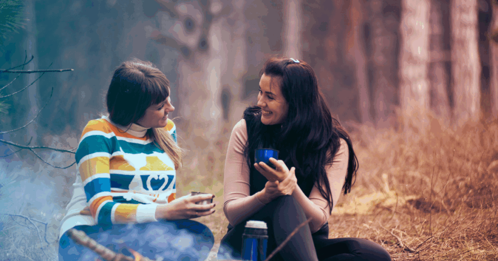 Two women enjoy a fireside chat in the woods, showcasing ambivert social behavior and outdoor lifestyle.